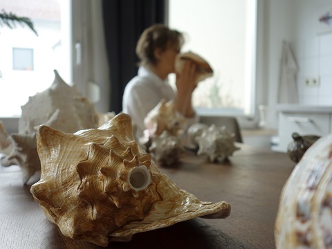 Künstlerin Kathleen Jahn bläst in ein Schneckenhorn, umgeben von einer Sammlung verschiedener Schneckenhörner auf einem Holztisch; im Hintergrund ein heller Wohnraum.
