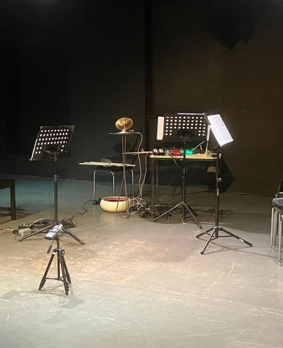 Bühne des ACUD-Theaters in Berlin mit Notenständern, Stühlen und elektronischen Musikinstrumenten.