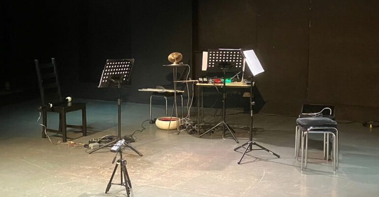 Bühne des ACUD-Theaters in Berlin mit Notenständern, Stühlen und elektronischen Musikinstrumenten.