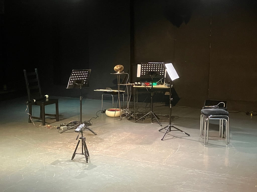Bühne des ACUD-Theaters in Berlin mit Notenständern, Stühlen und elektronischen Musikinstrumenten.
